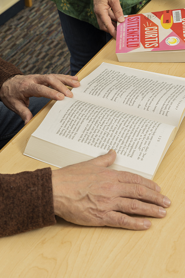 book on table adult hands on page with woman's hands on another book at left